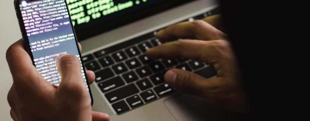 Free Unrecognizable hacker with smartphone typing on laptop at desk Stock Photo